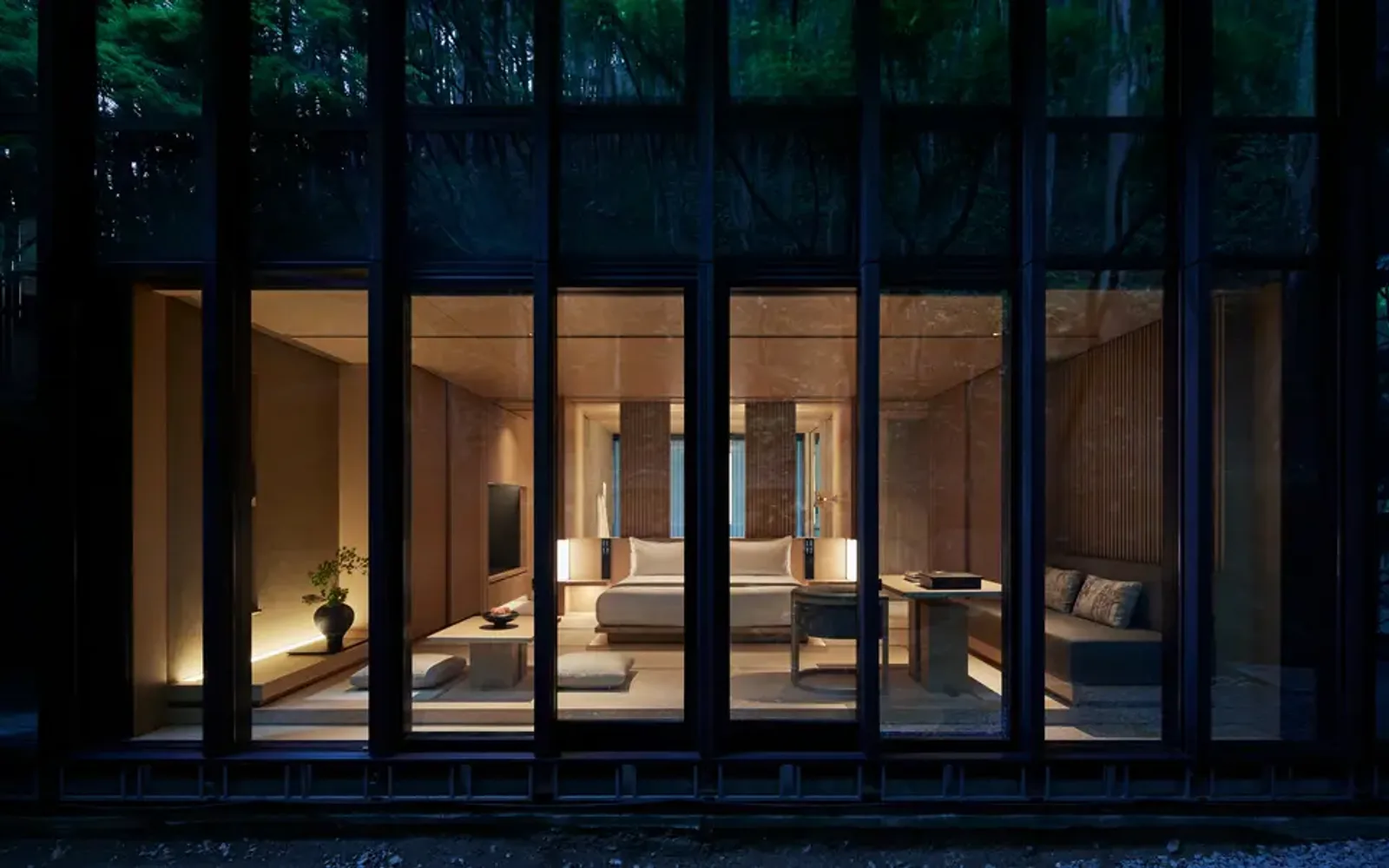 Pavilion bedroom at Aman Kyoto lit warmly at dusk, with floor-to-ceiling glass doors opening onto a dark courtyard.