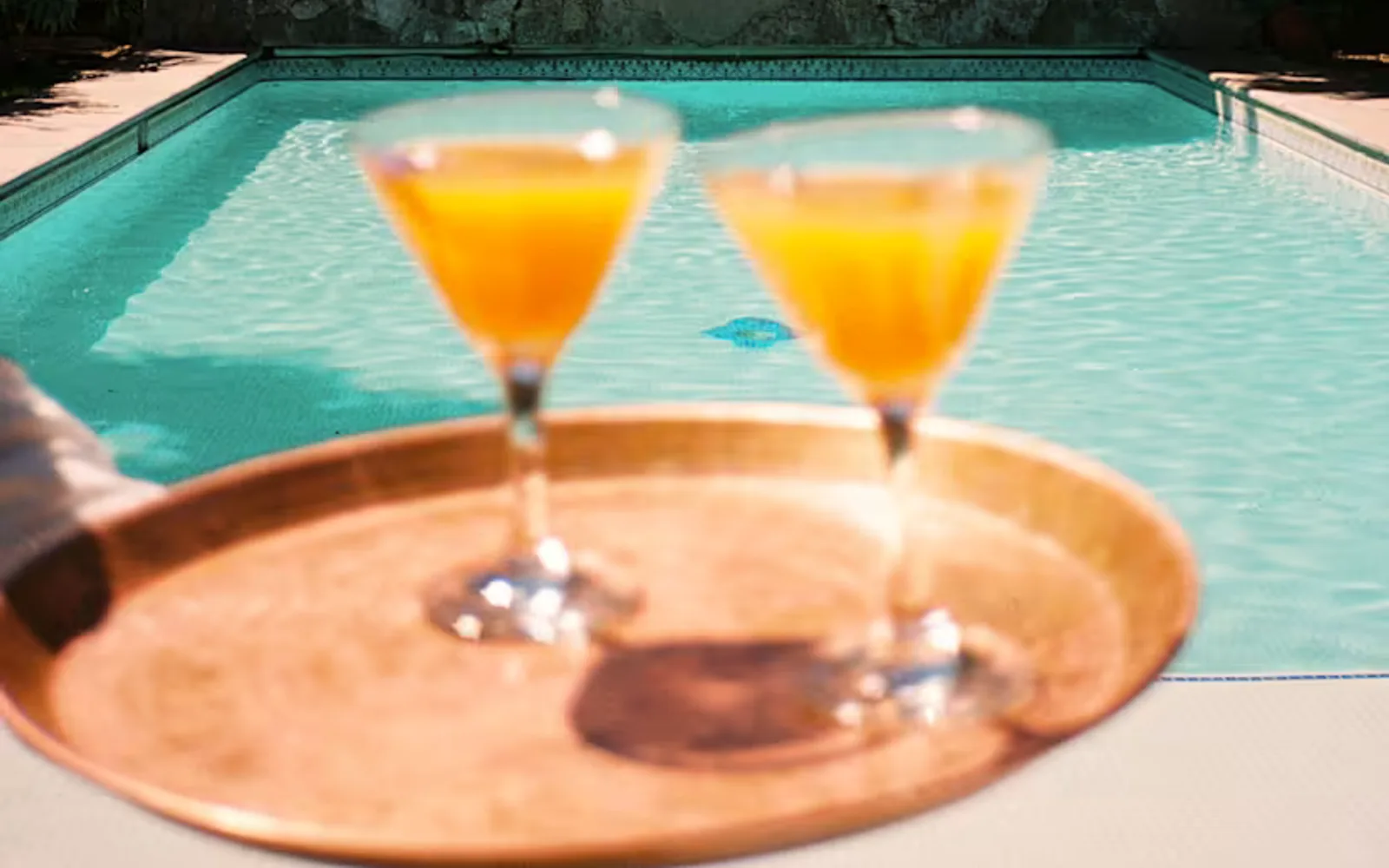 A waiter carries two orange drinks on a copper tray, seen close-up in soft-focus, with the defined sparkling pool behind.