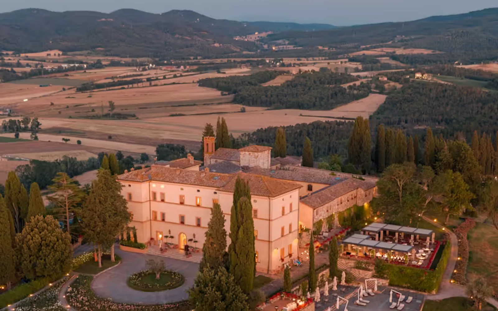 Magical aerial view of Castello di Casole at dusk, with the hotel, outdoor cinema and terrace illuminated with pretty lights.