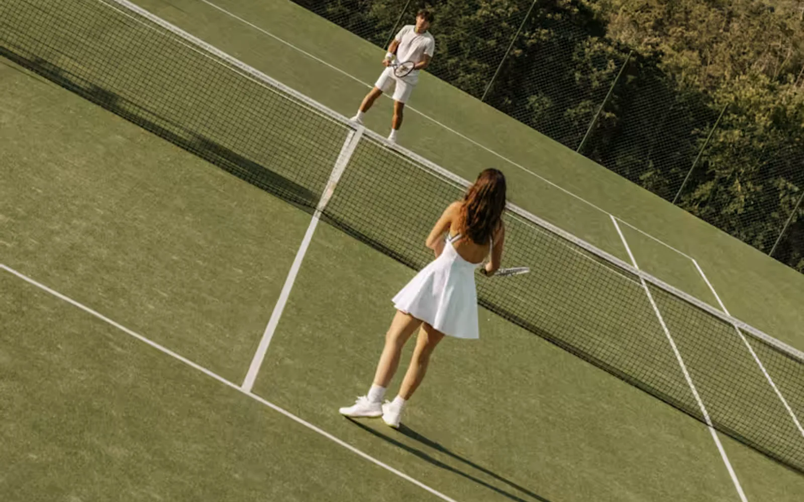 Angled image of a man and woman in tennis whites as they play on a grass court with forest views, seen from behind the woman.