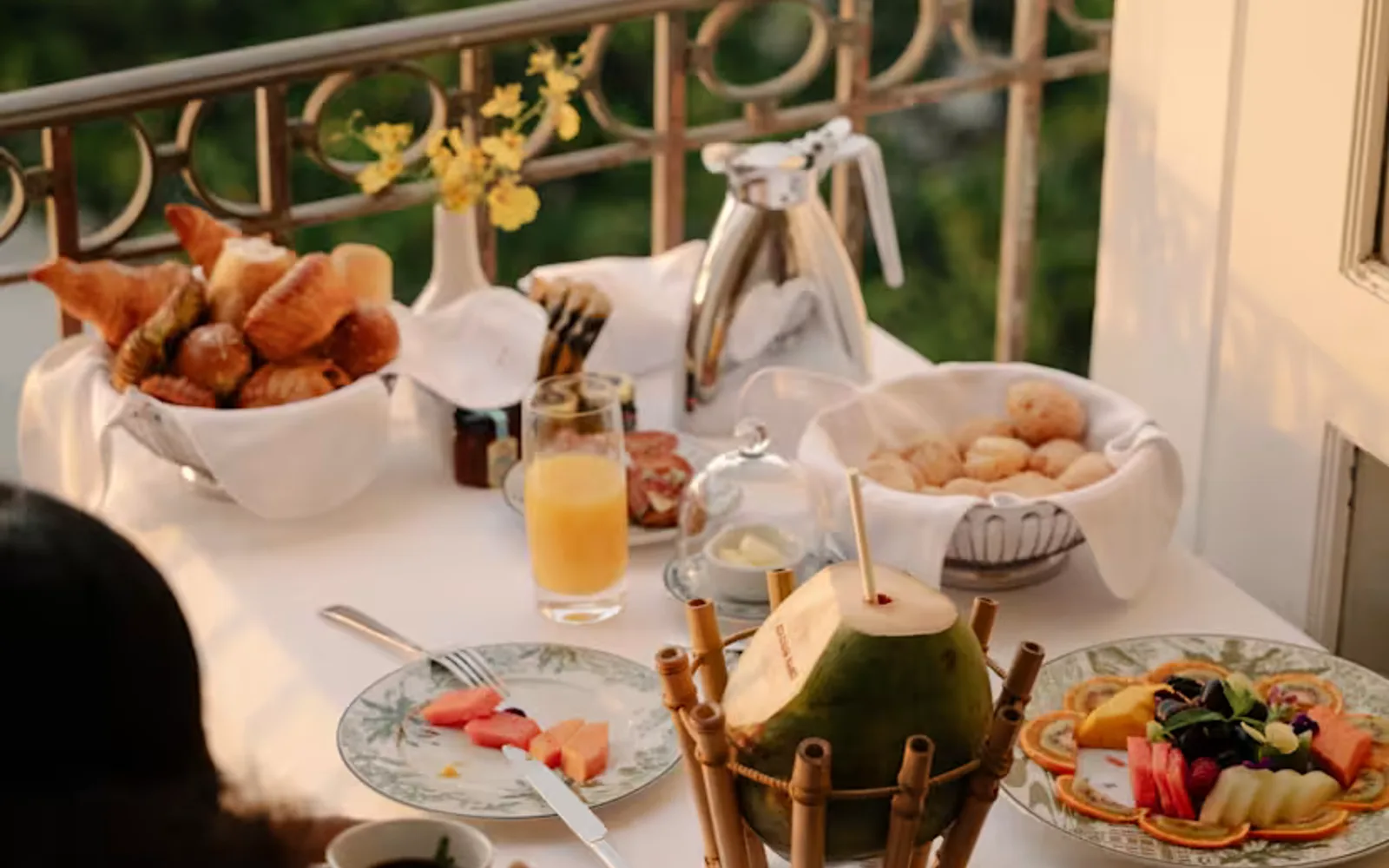 A table with fruit, pastries and a coconut with straw in a bamboo stand, seen over the shoulder of a woman dinking coffee.