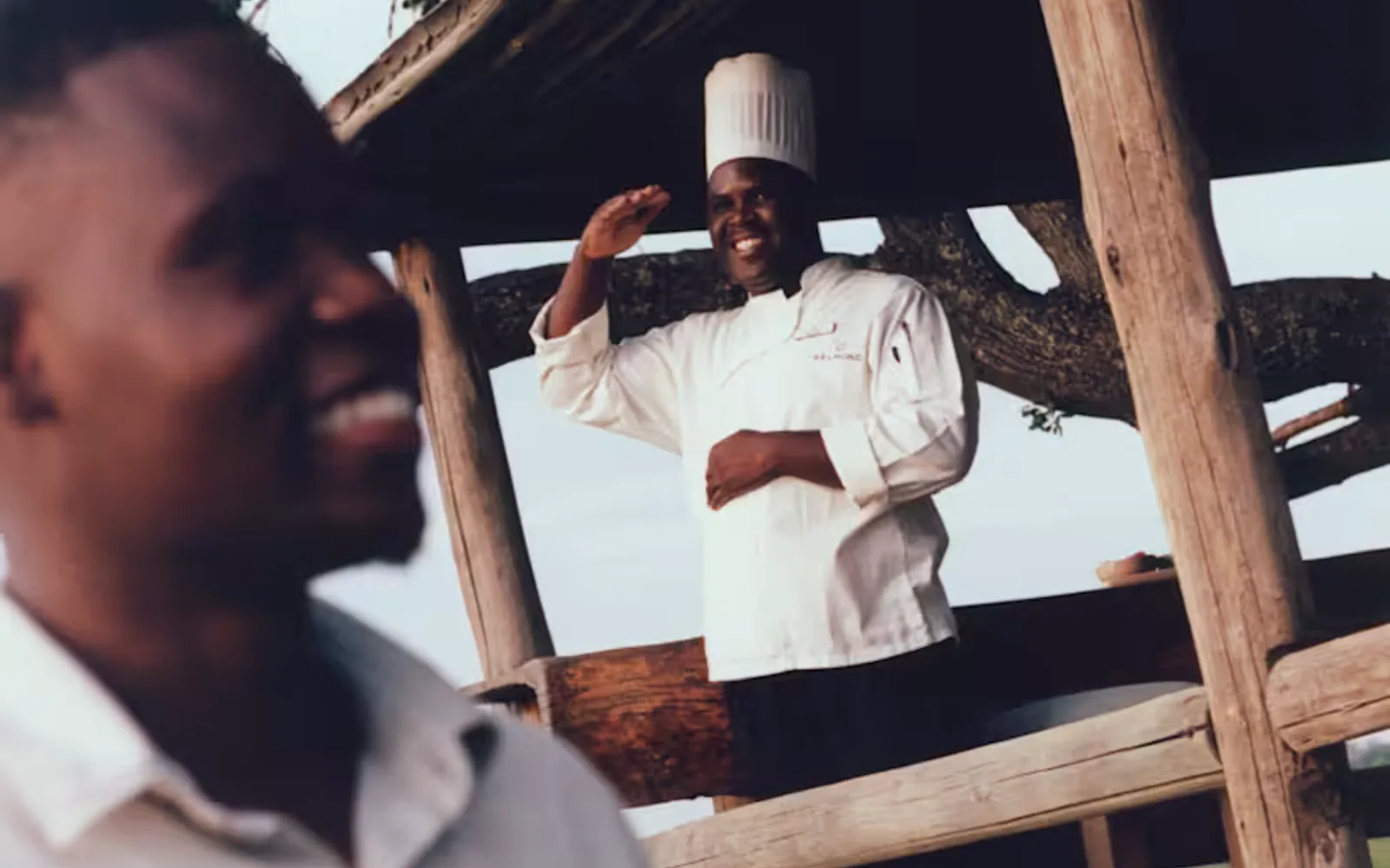 A chef in a toque smiles and salutes from the Fish-Eagle Bar, while a waiter with a tray passes by in soft-focus foreground.