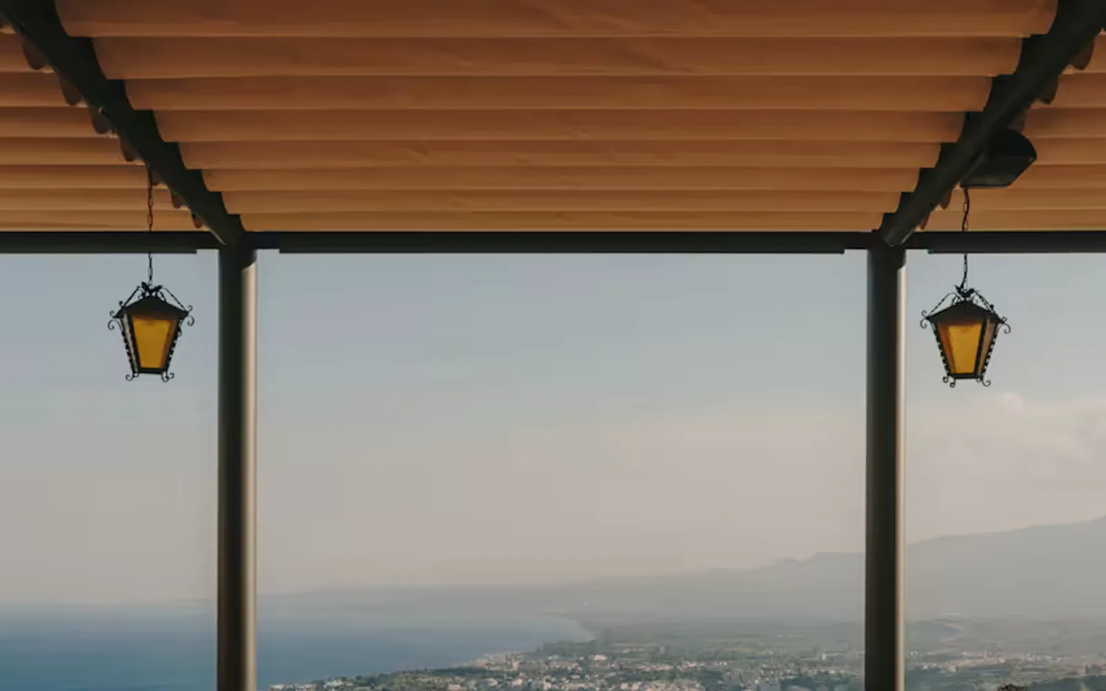 Laid tables with wicker chairs await guests on the Ristorante Timeo terrace, with a canopy and breathtaking coastline views.