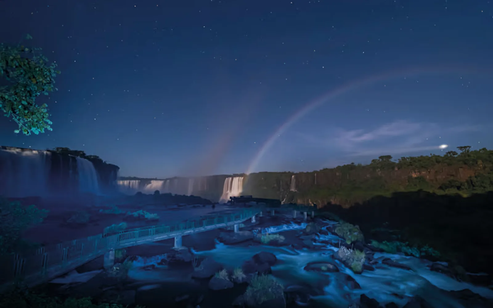 A faint moonbow rises from the cascading waters of the Iguassu Falls, seen at night over the phosphorous-blue river.