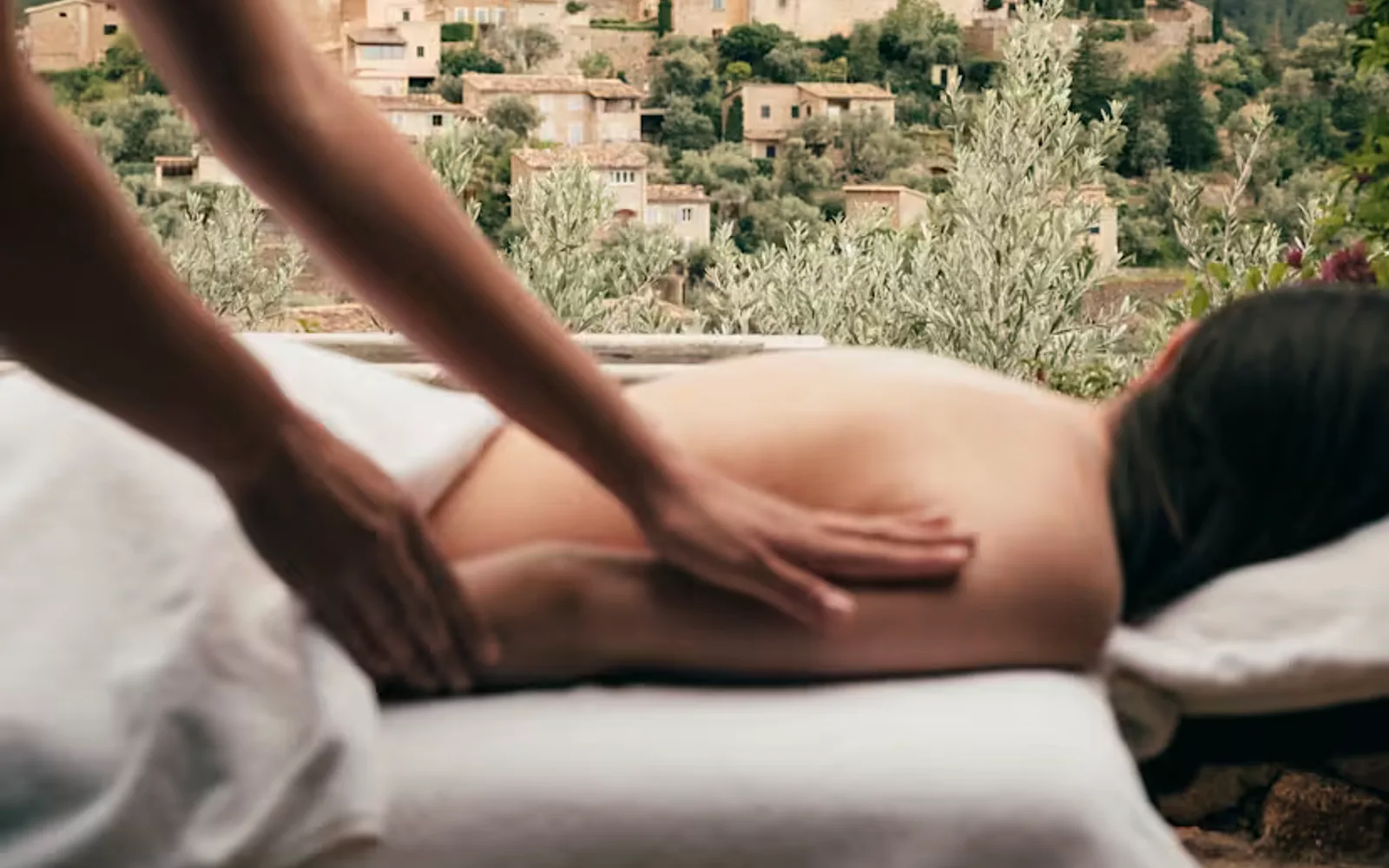 A therapist in soft-focus foreground performs a massage on a spa guest's back, with views behind of a pretty hilltop town.