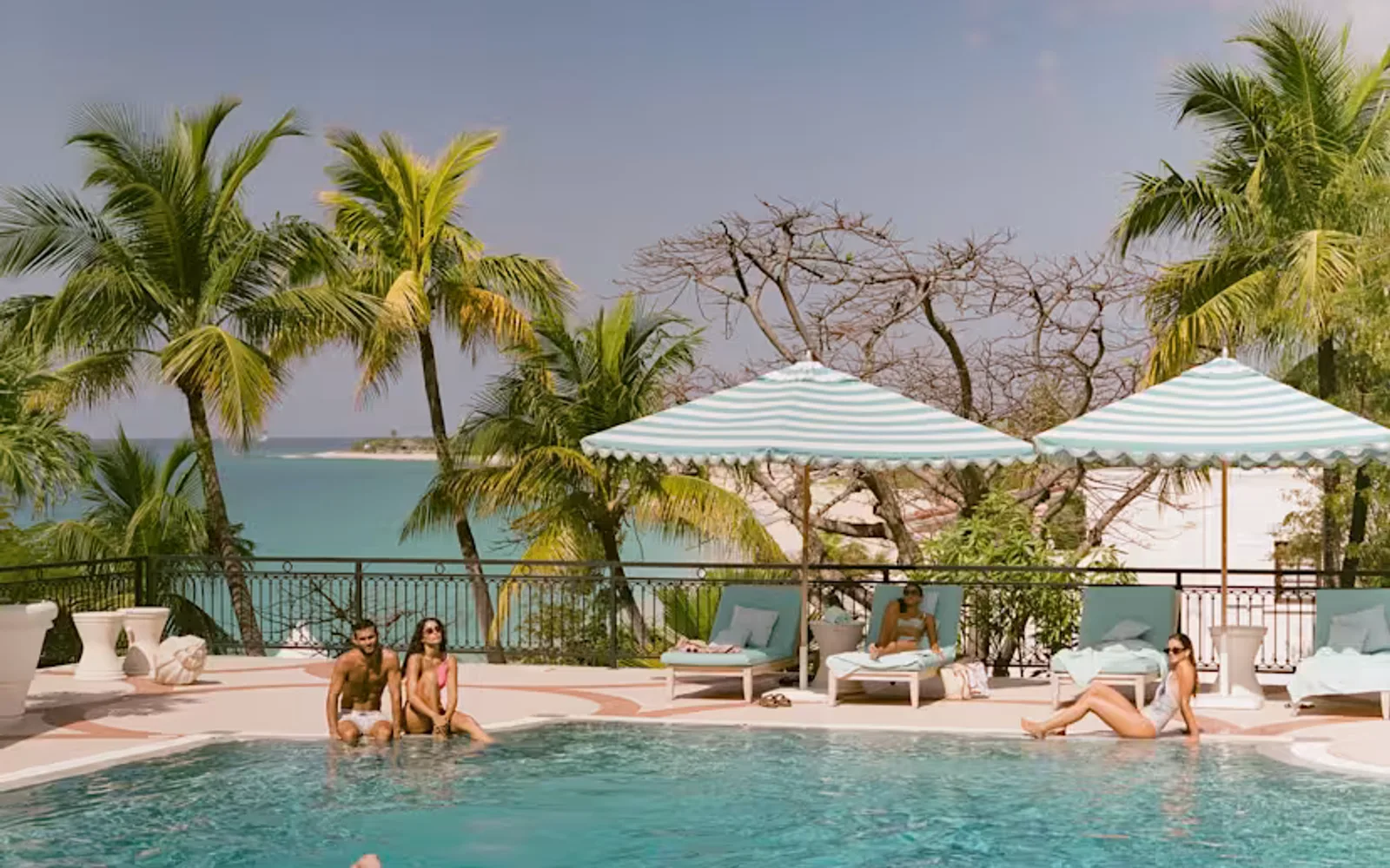 A man swims front-crawl across the pool, as three guests look on from the poolside, with a backdrop of palm trees and ocean.