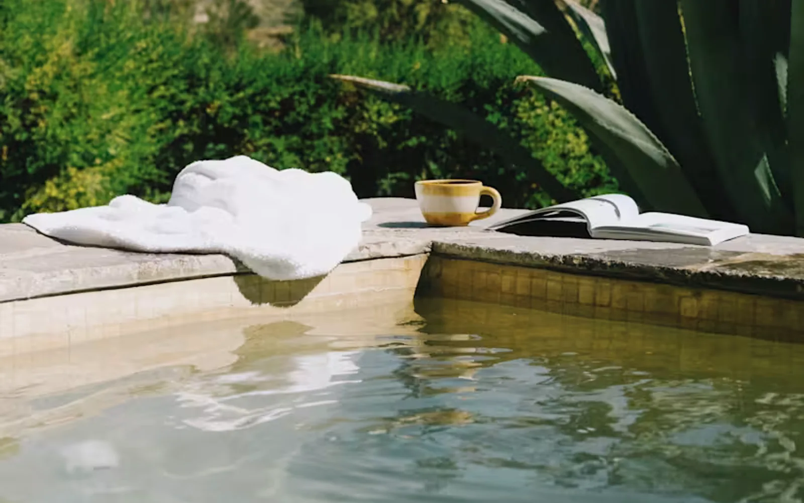 A white towel, mug and book rest on the corner edges of a pool in an eye-level view from the water, capturing hills beyond.