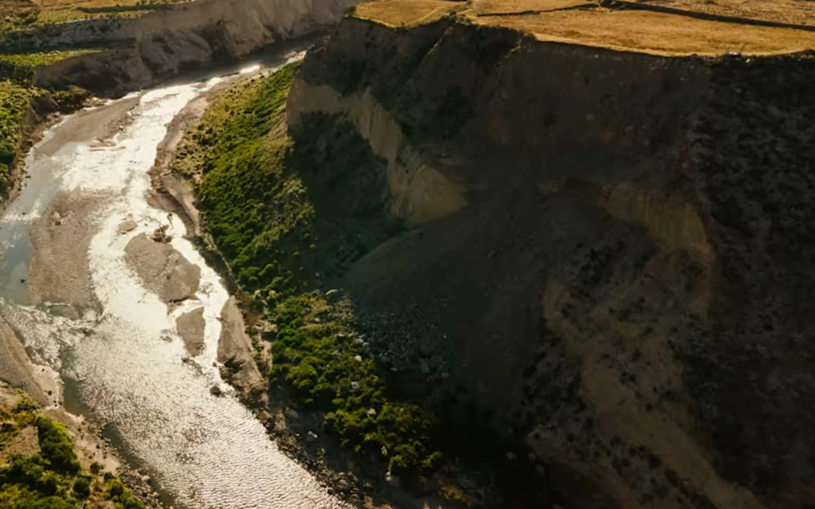 Sunlight glistens on the Colca river as it snakes between the green terraces and rock cliffs of the canyon, seen from above.