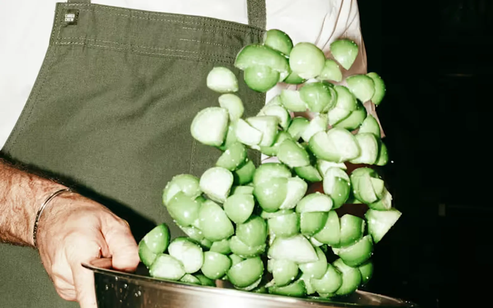 Chef Curtis Stone, with a short beard and dark-rimmed glasses, tosses chopped green tomatoes in a steel bowl, seen close-up.