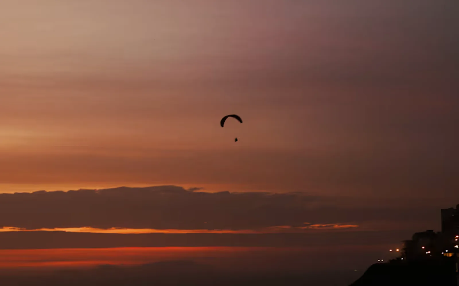 The hand-glider silhouetted against a crimson sunset sky soars above the trails of traffic lights along the Lima coastline.