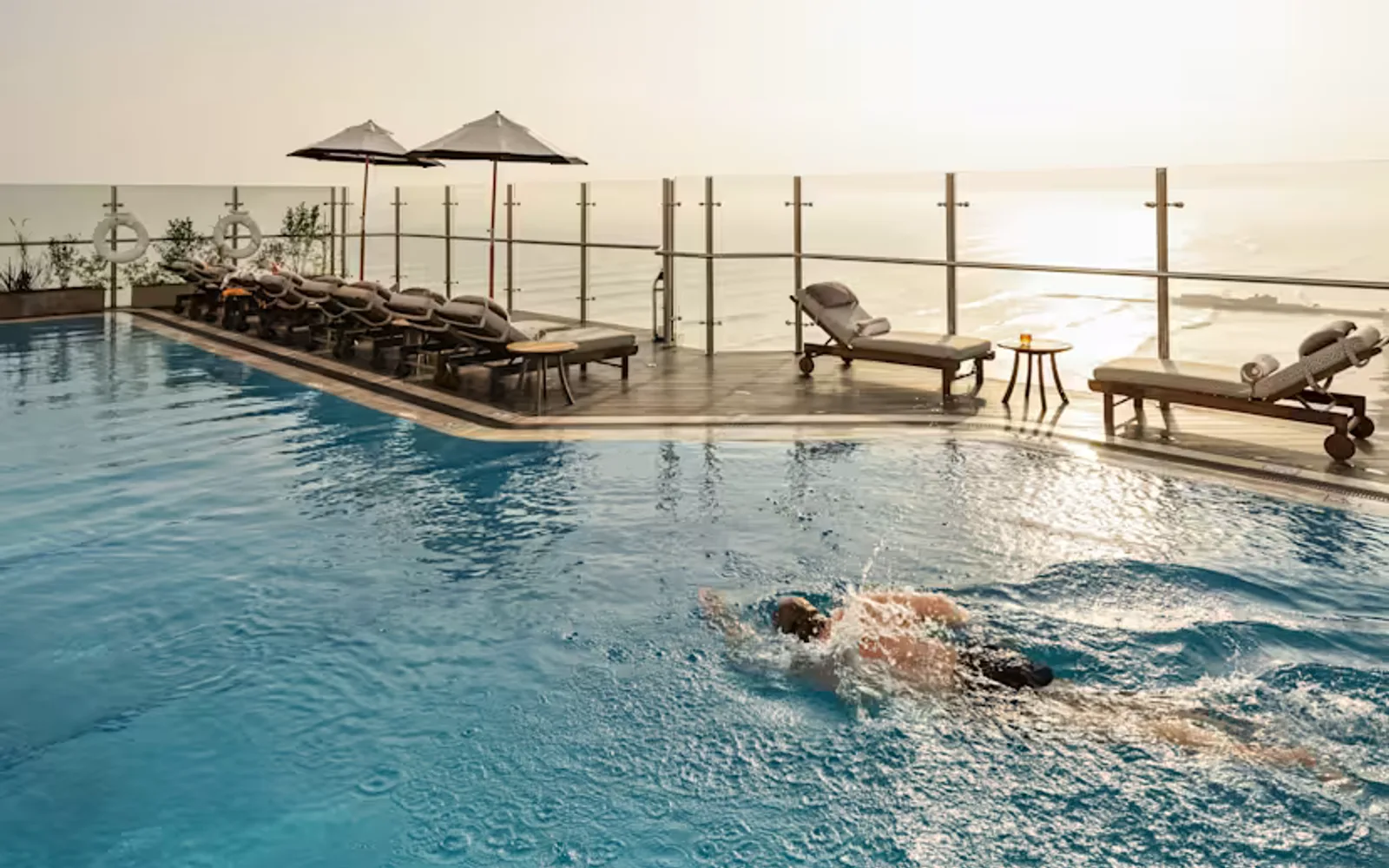 A man swims in the rooftop pool, in a view to the opposite side where eight loungers face away, towards the glowing ocean.