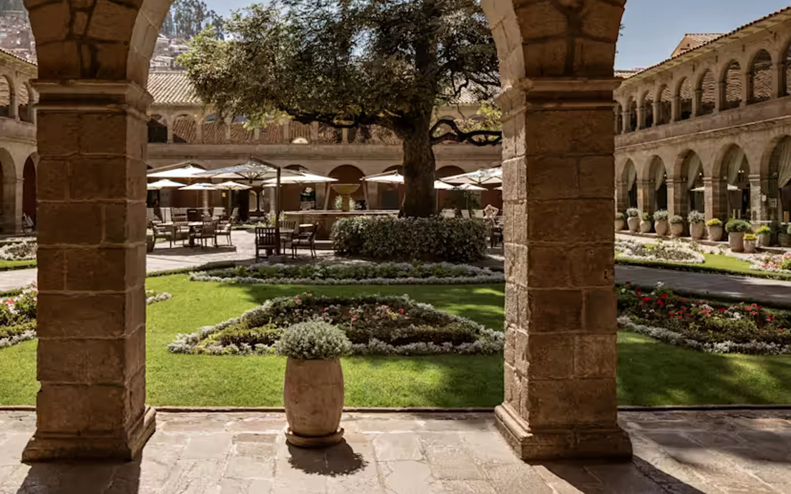 View of the courtyard with lush lawn sections, dining sets with parasols and a central cedar tree, viewed from the cloisters.