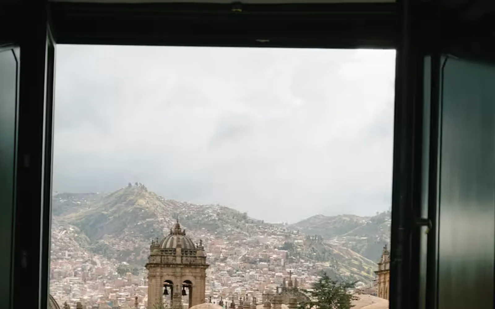 A woman sits at a table with coffee gazing out of her suite window at the terracotta roofscape of Cusco beneath a moody sky.