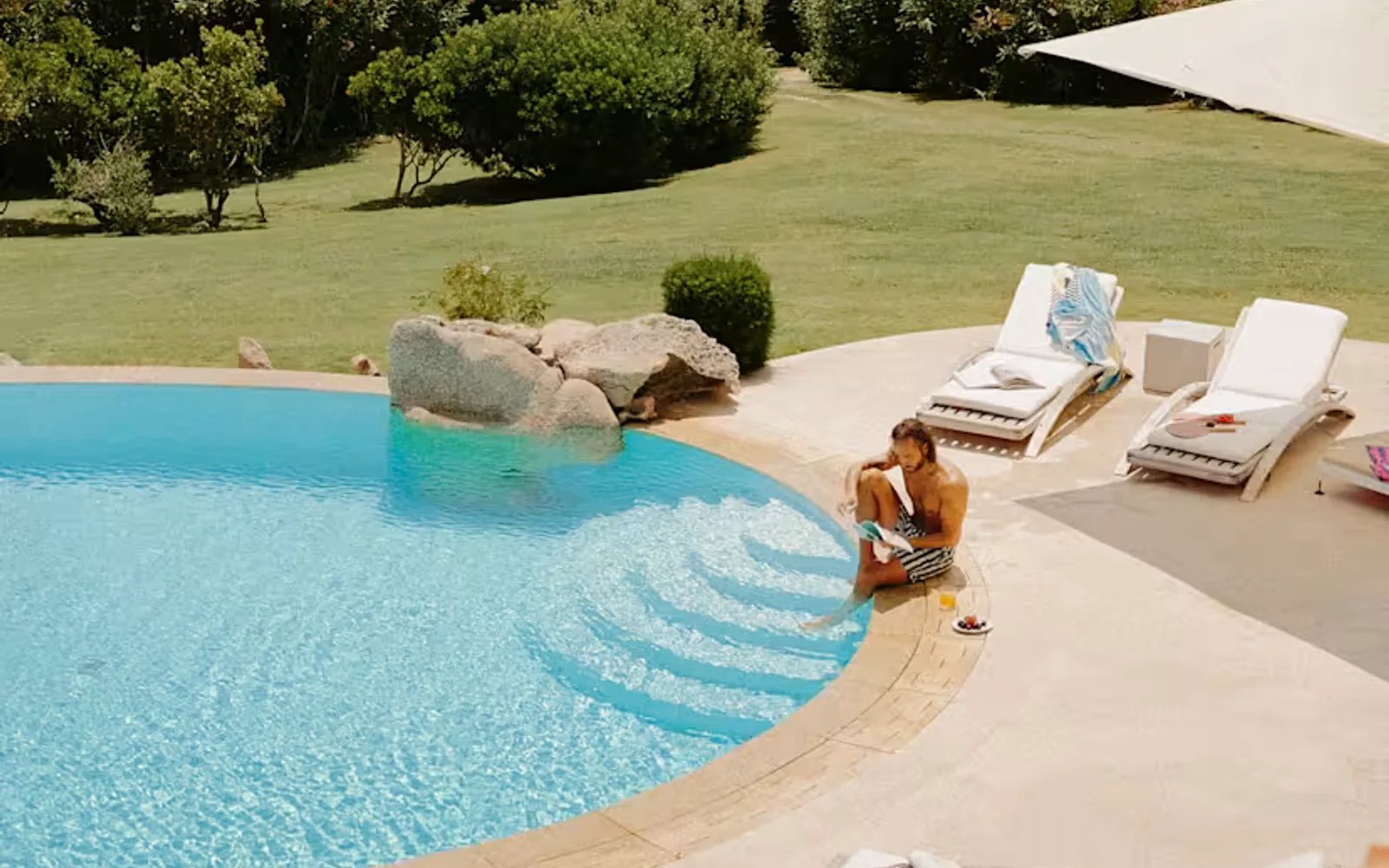 A man reads at the poolside of a villa pool with one foot in the water, seen in a high-angle view that captures the coast.