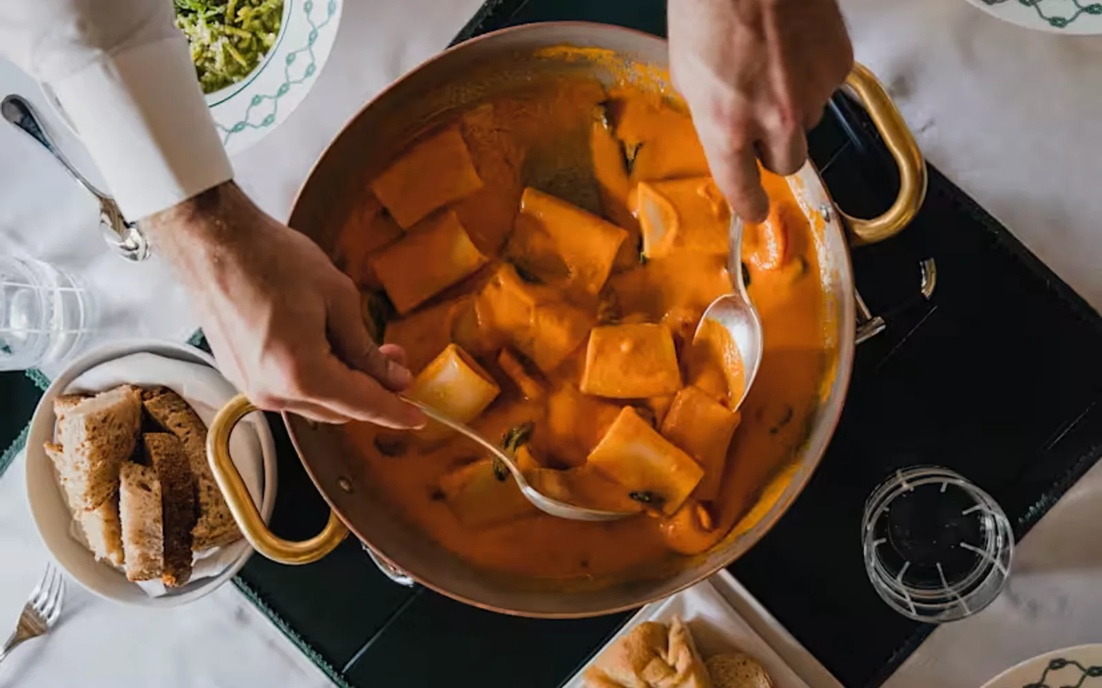 A man uses spoons to help himself to a portion of paccheri in rich, creamy tomato sauce, from a large pan, seen from above.
