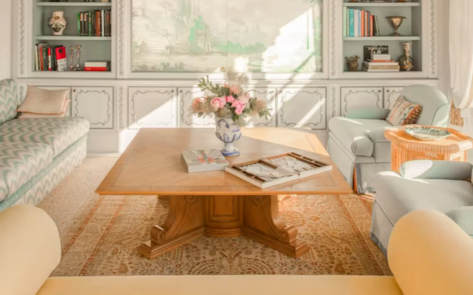 A bookcase, vintage sofa, two blue armchairs and yellow loveseat border a large coffee table beneath a beamed suite ceiling.