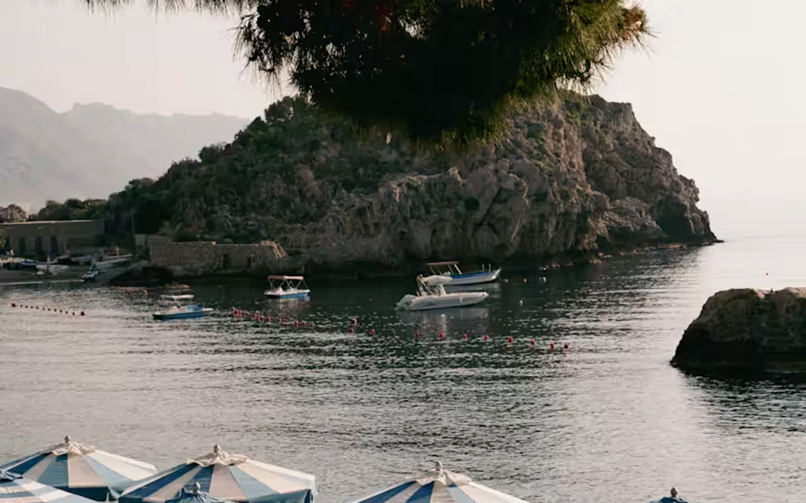 A shot of blue and white parasols on Mazzarò Bay beach at sundown, with views across still waters and boats to the headland.