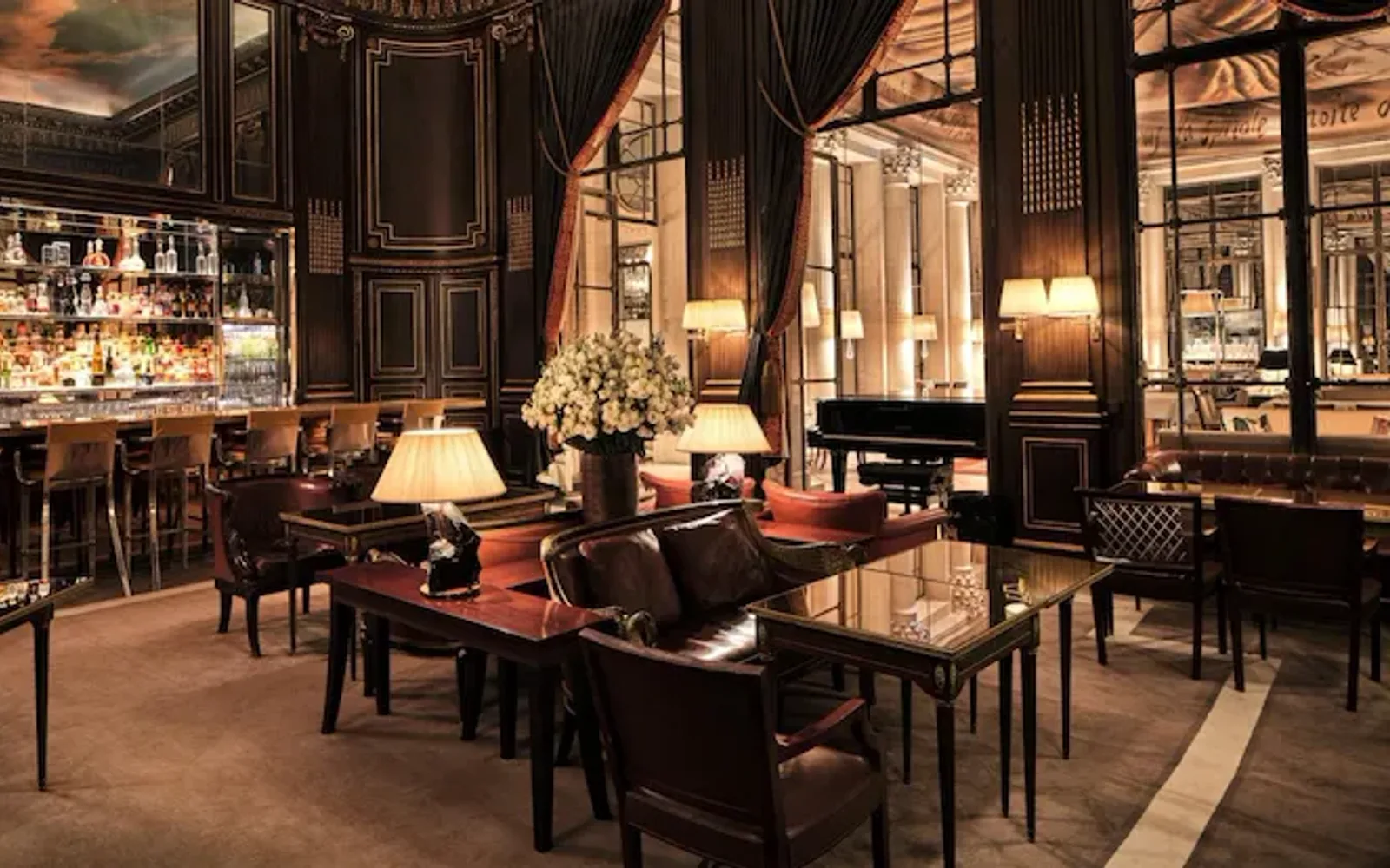 In the center of the photo, we see lamps lit on tables with armchairs and dark red cushions, and a bouquet of white flowers in a vase. On the left is bar 228 with high chairs. We can see the bottles on the shelves. Above the bar is a mirror reflecting the ceiling, which represents a sky with clouds. We look at the woodwork on the walls, with its gilded details. To the right is a piano and large windows with red curtains, revealing another room at Le Meurice, Paris.