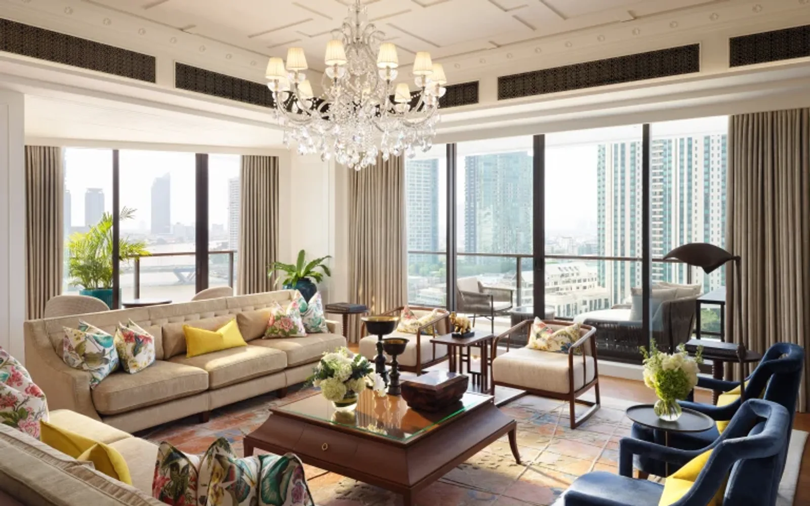 The Oriental Suite living room with a crystal chandelier, floral cushions, navy armchairs, and panoramic views of the Bangkok skyline and Chao Phraya River.