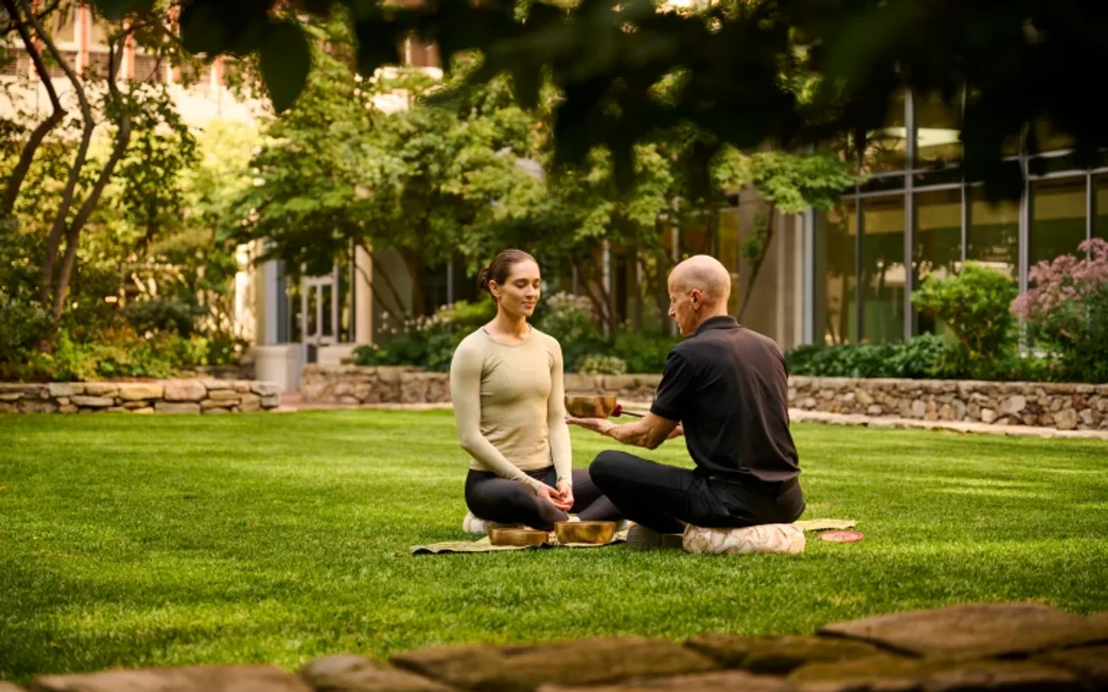 A wellness practitioner and a guest sit outdoors in garden terrace facing each other during a Himalayan singing bowl session.