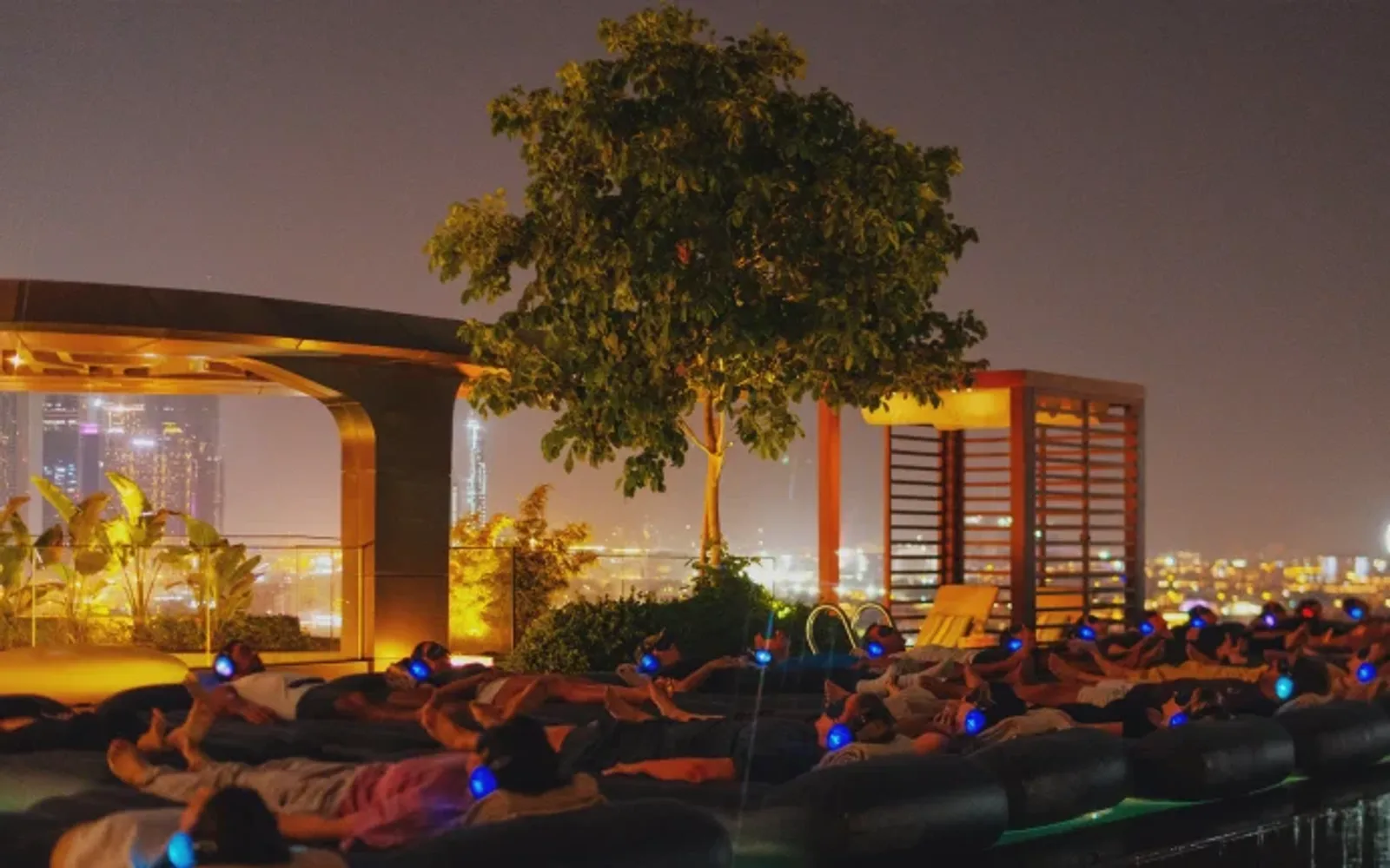 People lounging on pool floats overlooking a cityscape at night, under a large tree.