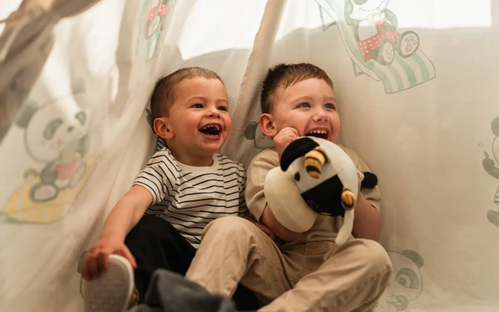Two young children playing inside a fabric tent, holding a stuffed toy.