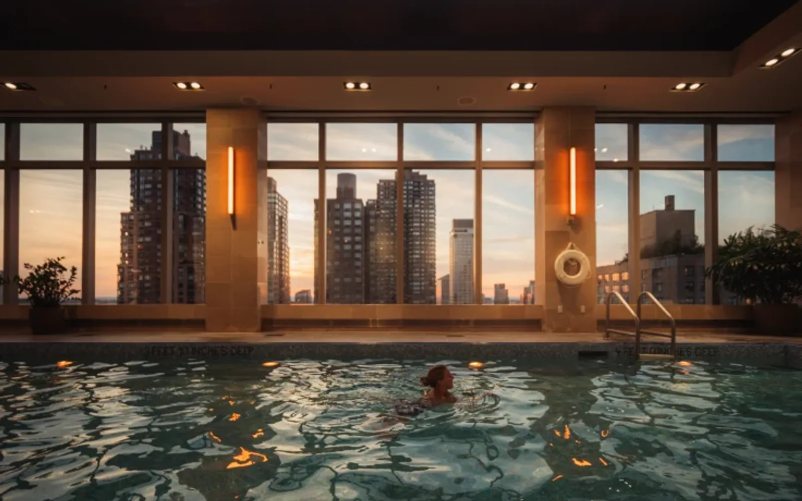 A person swims in an indoor pool with windows letting in sunset light and a view of buildings.