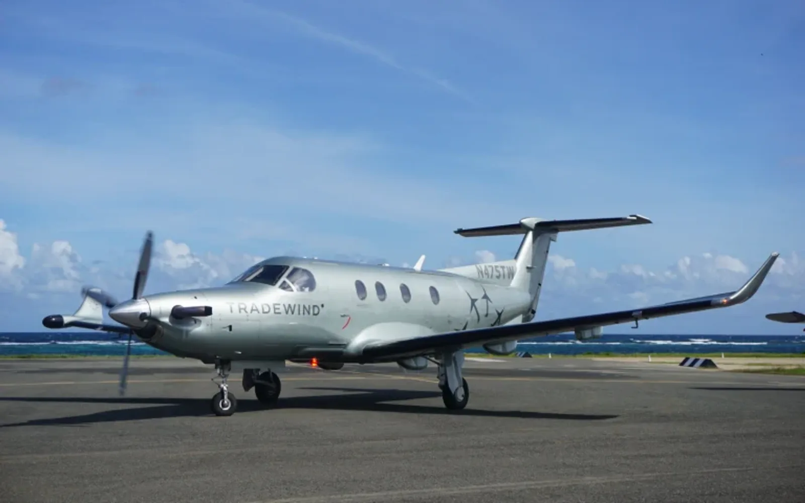 Silver Tradewind airplane parked on an airport runway near the ocean, with propellers spinning and a blue sky with scattered clouds in the background.