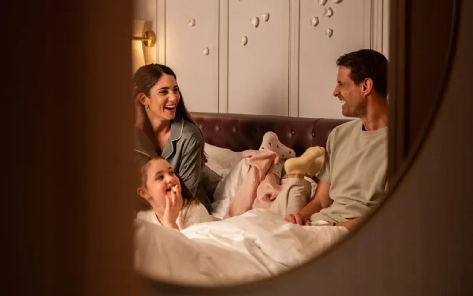 A warm family moment in a comfortable hotel room, with parents and child relaxing together on the bed and sharing laughter.
