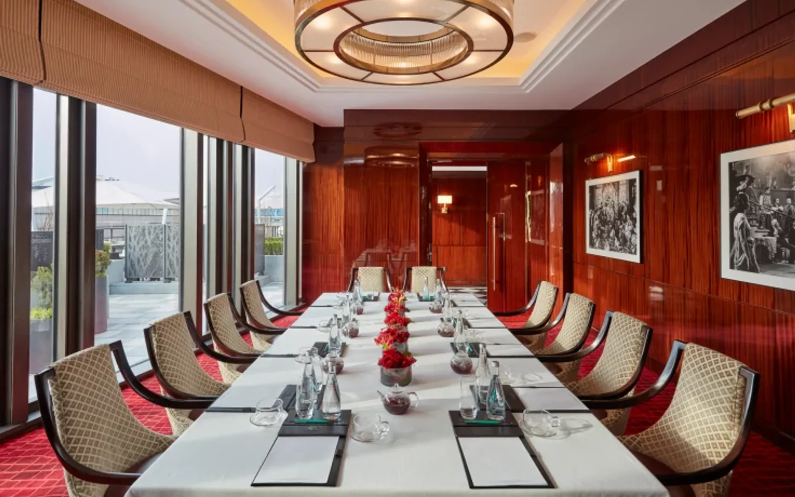 A meeting room features a long, set table with white linens and red florals, patterned chairs, rich wood paneling, a sleek circular chandelier, large windows, and framed black-and-white art, radiating sophisticated refinement at Mandarin Oriental Wangfujing, Beijing.